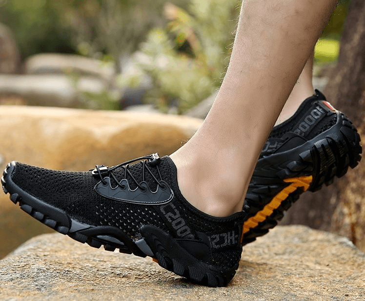 Breathable elastic men's camping sneakers in black with mesh upper and rubber outsole, shown outdoors on rocky terrain.